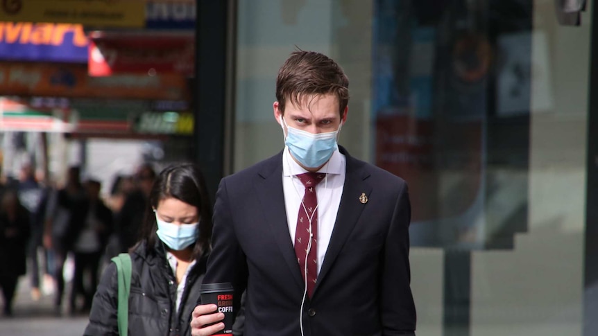 A man in a suit crosses the road while wearing a surgical mask, and holding a morning coffee