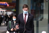 A man in a suit crosses the road while wearing a surgical mask, and holding a morning coffee