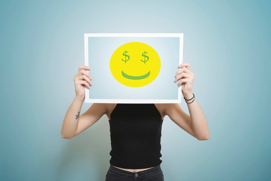 A person holds up a sign with a smiley face on it and dollar signs for eyes in front of their face.
