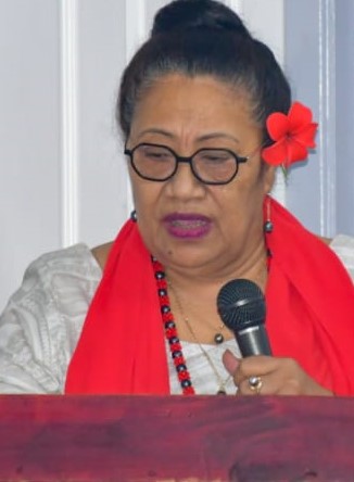   A woman with hair tied back wears a red flower above her ear as she speaks into a microphone