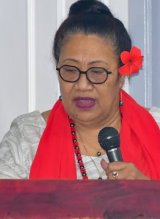  A woman with hair tied back wears a red flower above her ear as she speaks into a microphone