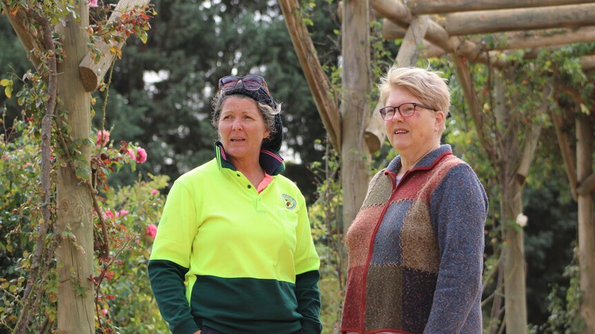 a woman wearing a hi-vis shirt speaks with another woman in a rose garden in a wooden walkway