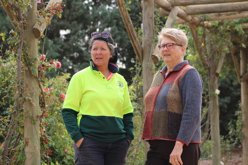 a woman wearing a hi-vis shirt speaks with another woman in a rose garden in a wooden walkway
