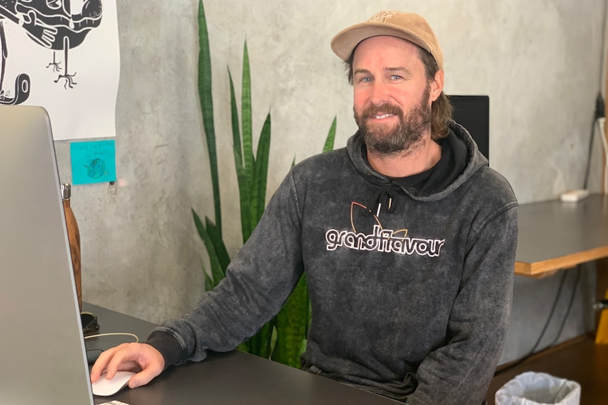 A man in a hoodie and hat sitting at a desk with a computer in front of him.