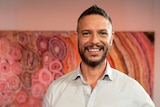 An Aboriginal man with short hair, beard and white shirt standing in front a colourful Aboriginal dot painting
