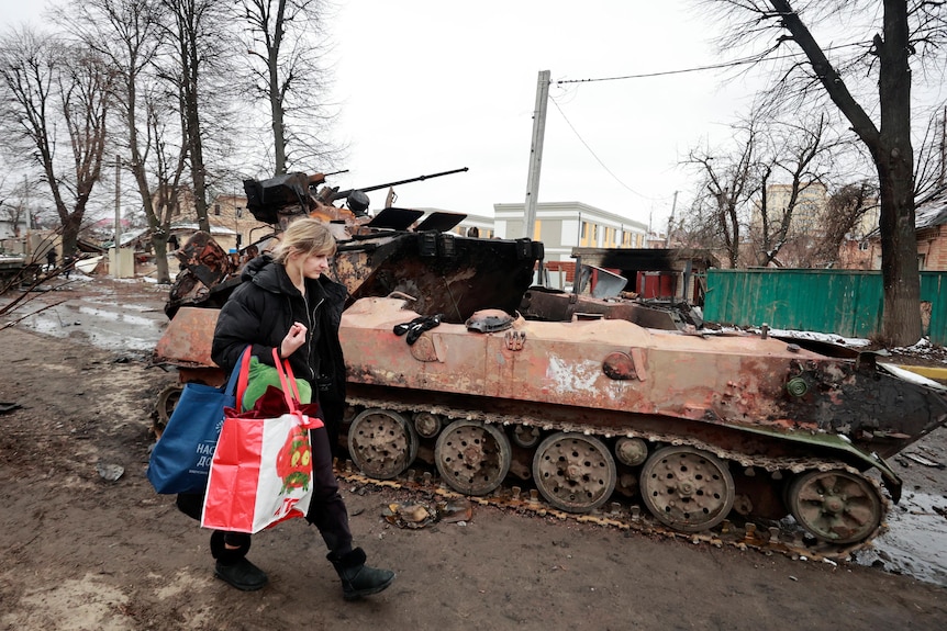 A woman carries shopping bags as she walks passed a burned tank