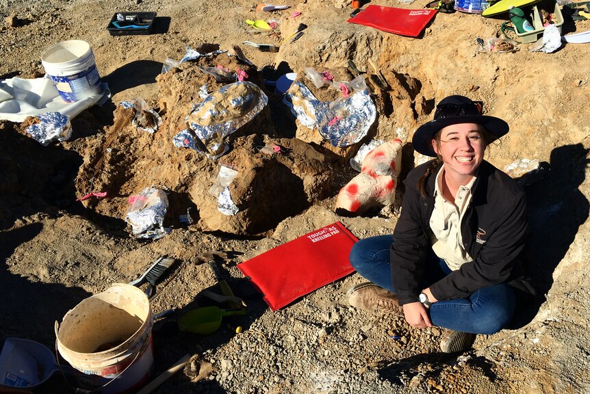 A woman sits on the ground. There is a hole next to her and digging equipment. She is smiling