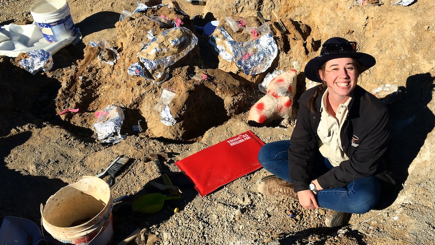 A woman sits on the ground. There is a hole next to her and digging equipment. She is smiling