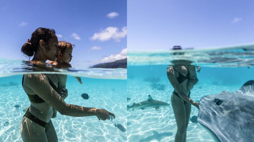 Woman holds young child standing in blue clear ocean, surrounded by fish and stingray