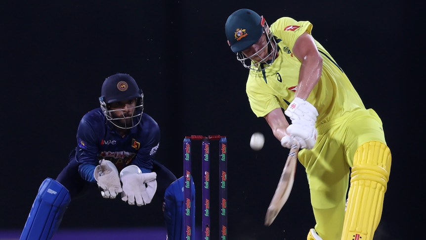 A cricket player wearing yellow hits a ball during a match while a wicket keeper wearing blue watches on