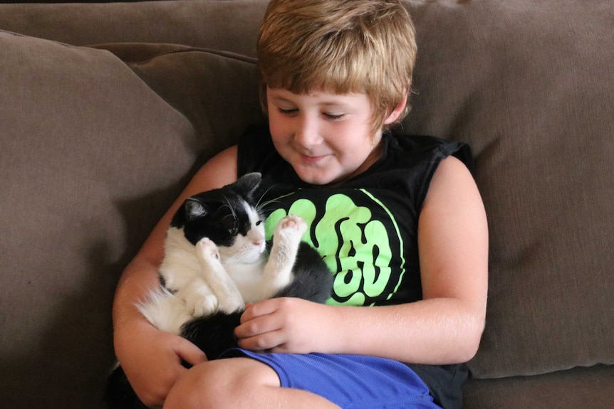 Seven-year-old Jett Thorne smiles as he sits on a brown couch holding his family's pet cat.