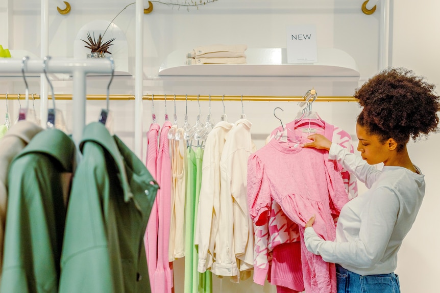 A woman holds up a pink dress and looks at it thoughtfully in a clothing store.