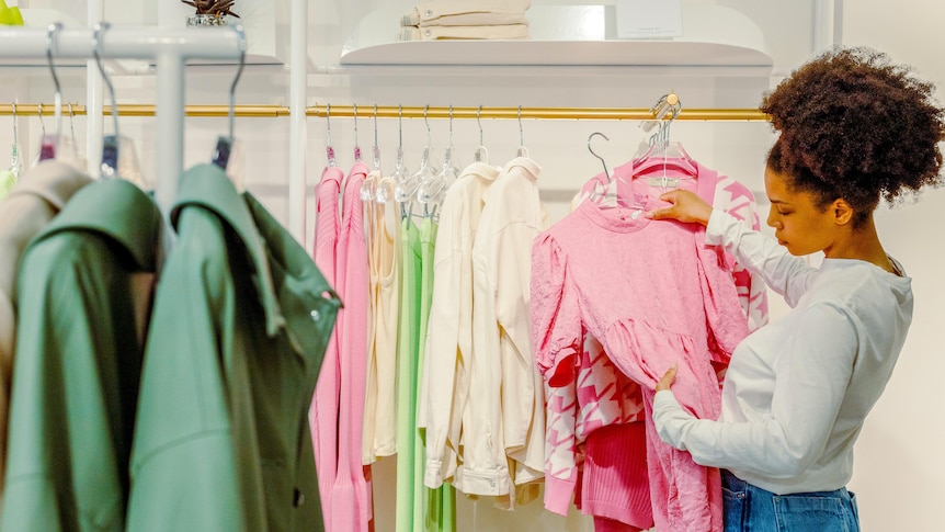 A woman holds up a pink dress and looks at it thoughtfully in a clothing store.