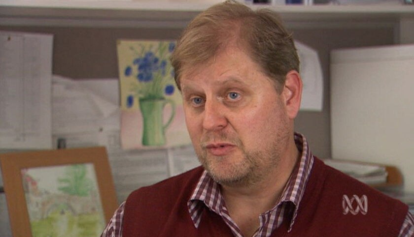 A bearded, brown-haired man in a maroon, checked shirt and maroon knitted vest in an office environment