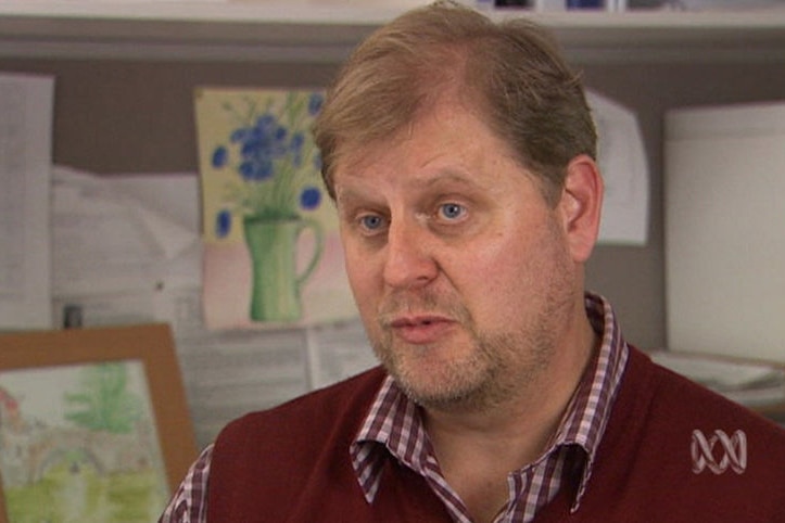 A bearded, brown-haired man in a maroon, checked shirt and maroon knitted vest in an office environment