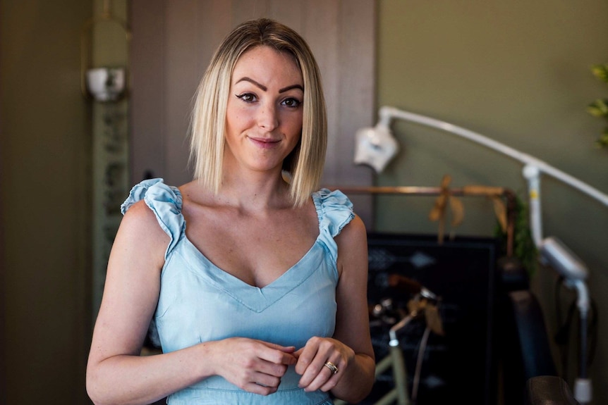 A young woman wearing a blue top stands posing for a photo and smiling.