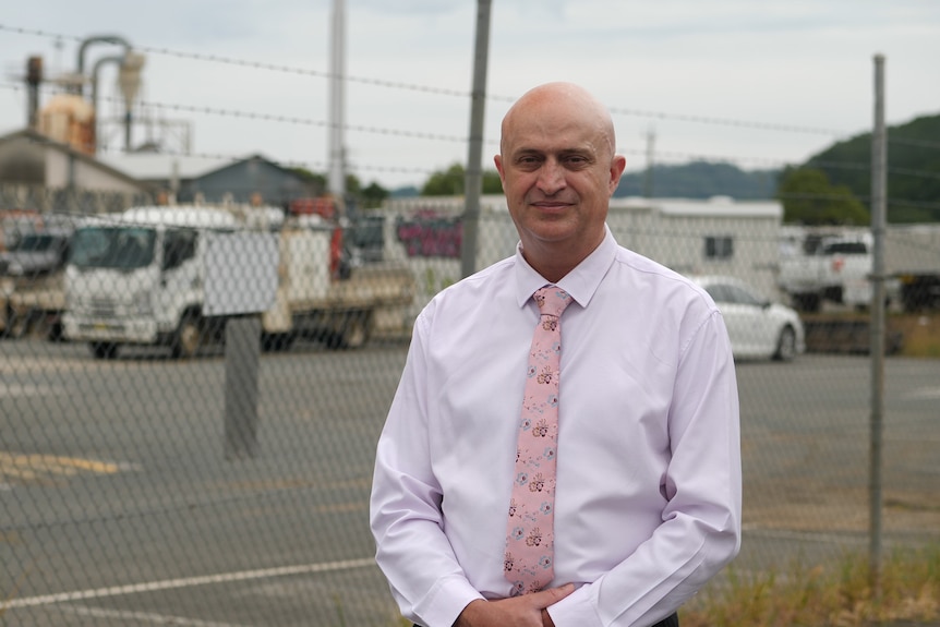 A man in a white business shirt in front of a wire fence with trucks and machinery in the background.