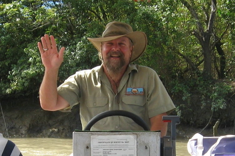 A man in a khaki shirt and hat waves from the wheel of a river cruiser