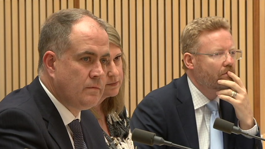 Two men and a woman wearing formal clothing sit in front of microphones in a wood panelled room.