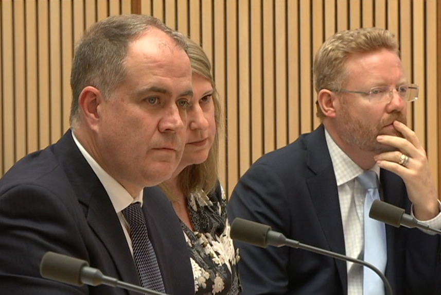 Two men and a woman wearing formal clothing sit in front of microphones in a wood panelled room.