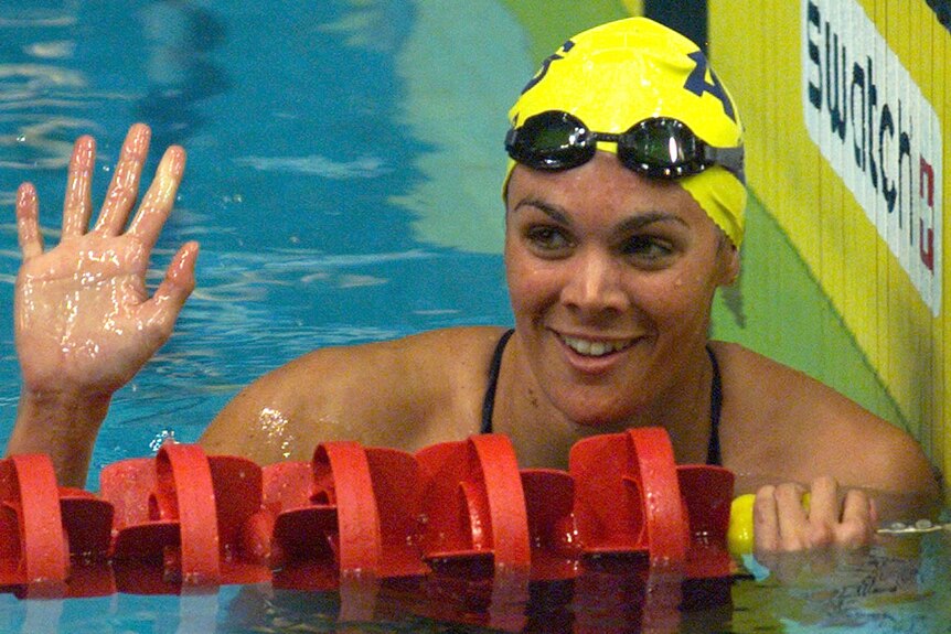 Samantha Riley wears a yellow swimming cap and waves from a pool lane.