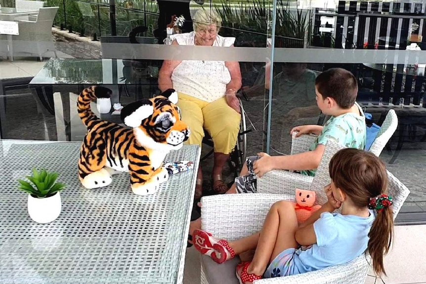 Lorraine Archer sits inside with her two grandchildren talking to her through glass at her aged care home.