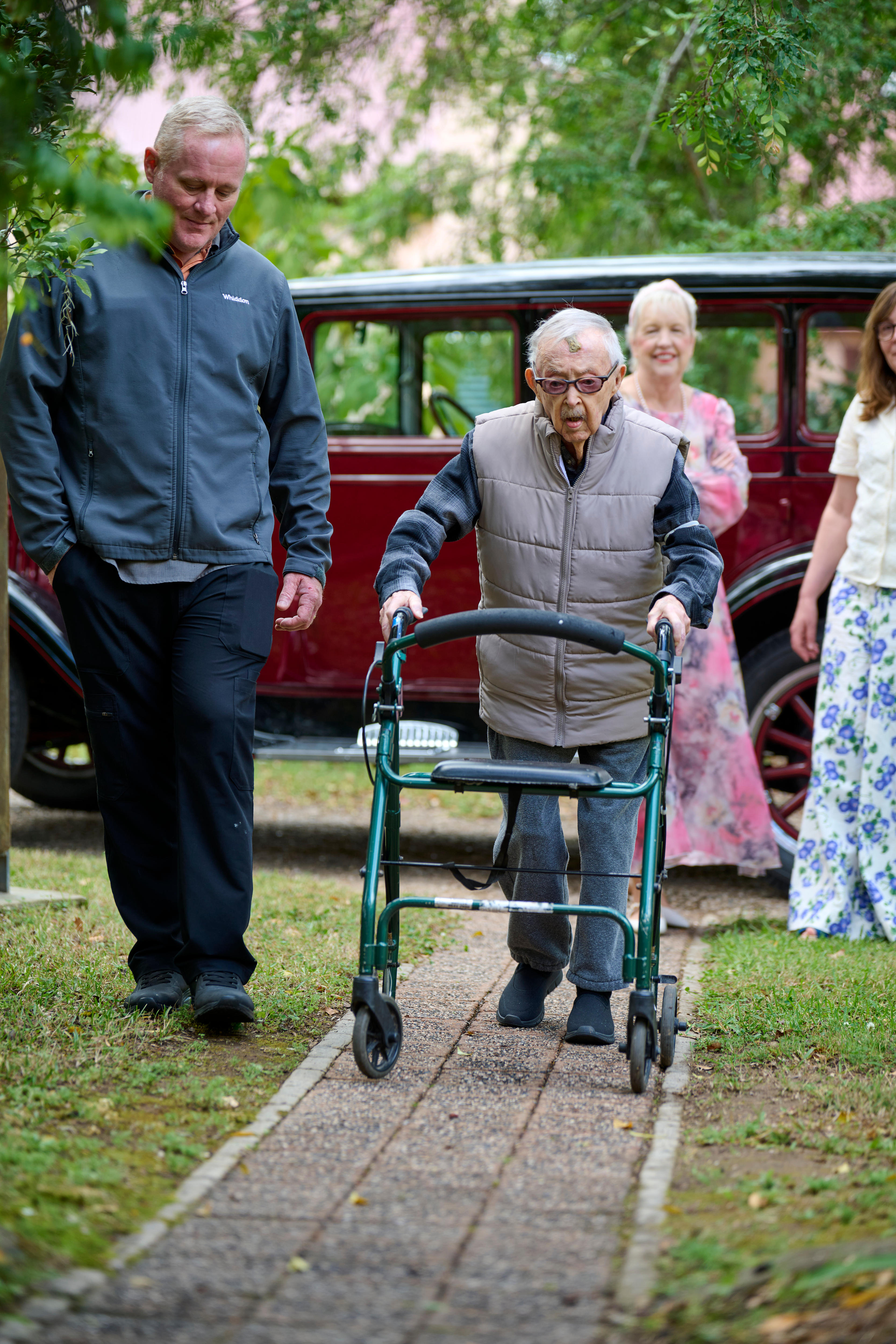Ken Weeks walks with a walking frame along a path. People in the background.