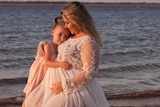 A woman and a small girl wearing dresses embrace on the beach