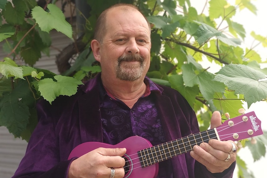Richard Tonkin stands outside holding a ukulele, he is wearing a purple velvet jacket and shirt.