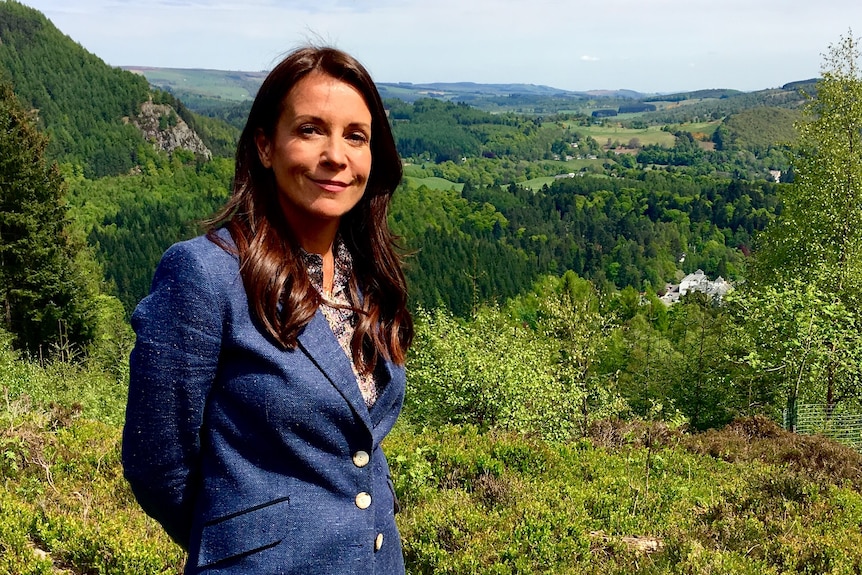 A woman in her early 40s with long brown hair and wearing a blue blazer stands in lush green countryside