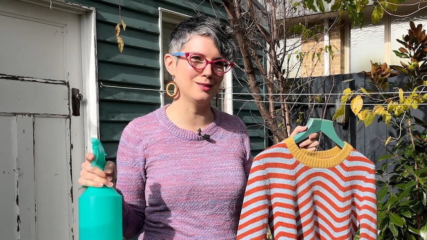 Erin Lewis-Fitzgerald holding a spray bottle in one hand, and stripy jumper in the other in front of garden shed and tree