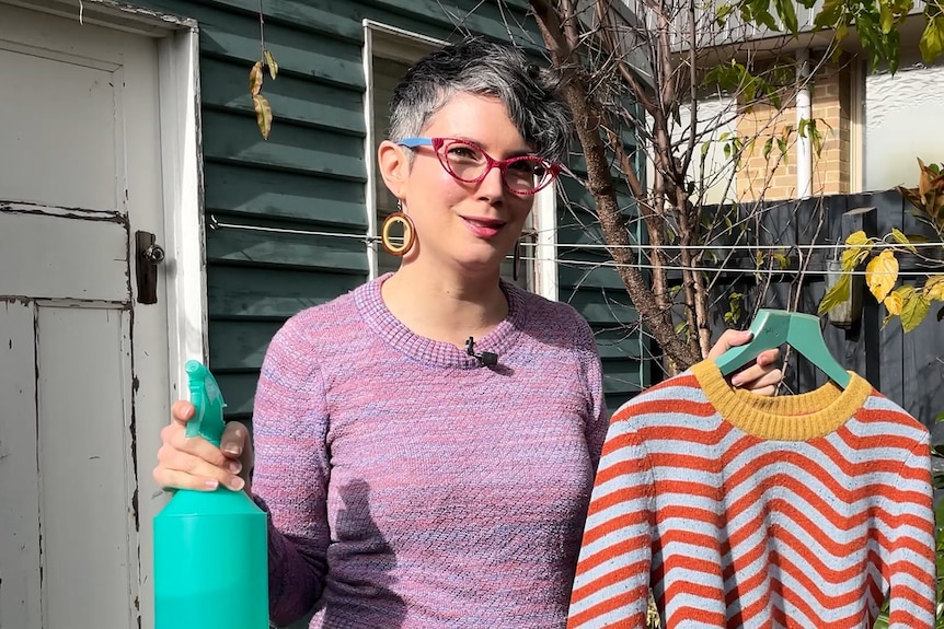 Erin Lewis-Fitzgerald holding a spray bottle in one hand, and stripy jumper in the other in front of garden shed and tree