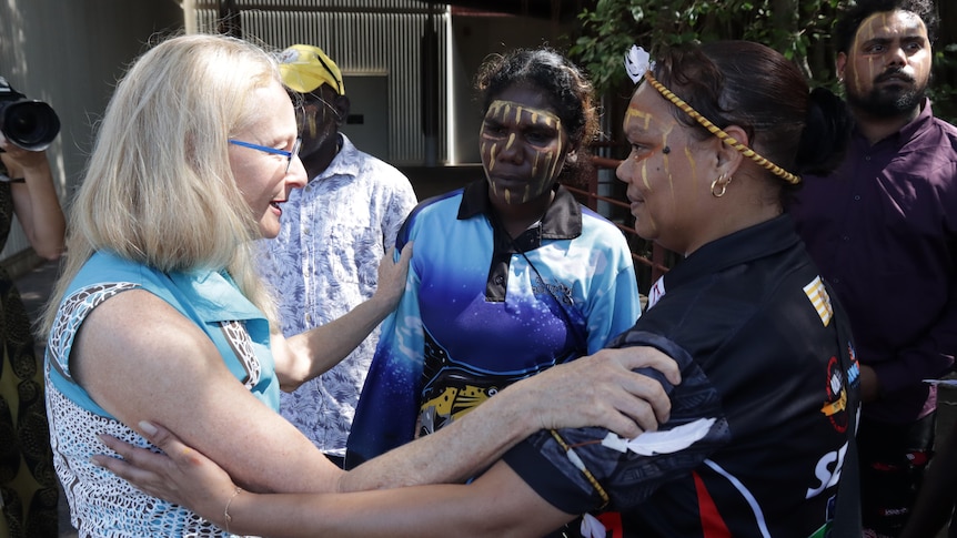 a blonde woman wearing glasses touches the shoulders of two aboriginal women wearing traditional face paint