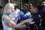 a blonde woman wearing glasses touches the shoulders of two aboriginal women wearing traditional face paint