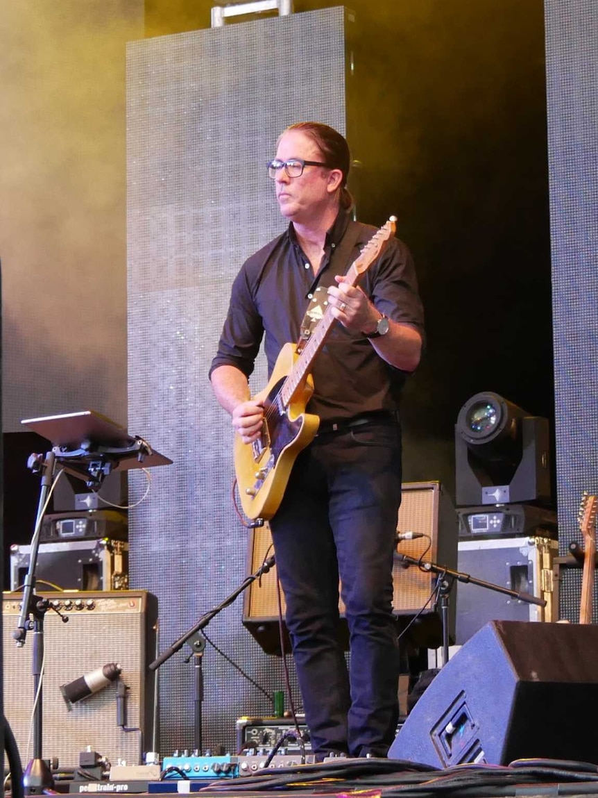 A man dressed in black stands on a stage playing guitar