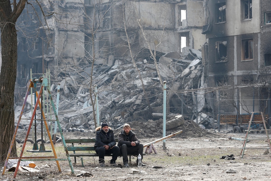 Two men in coats sit on a bench while in the background buildings are crumbling.