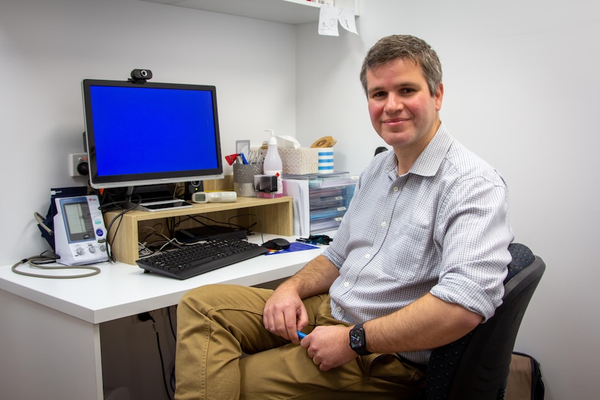 A GP in a blue shirt and beige pants looks towards the camera, while sitting at a desk with a computer 