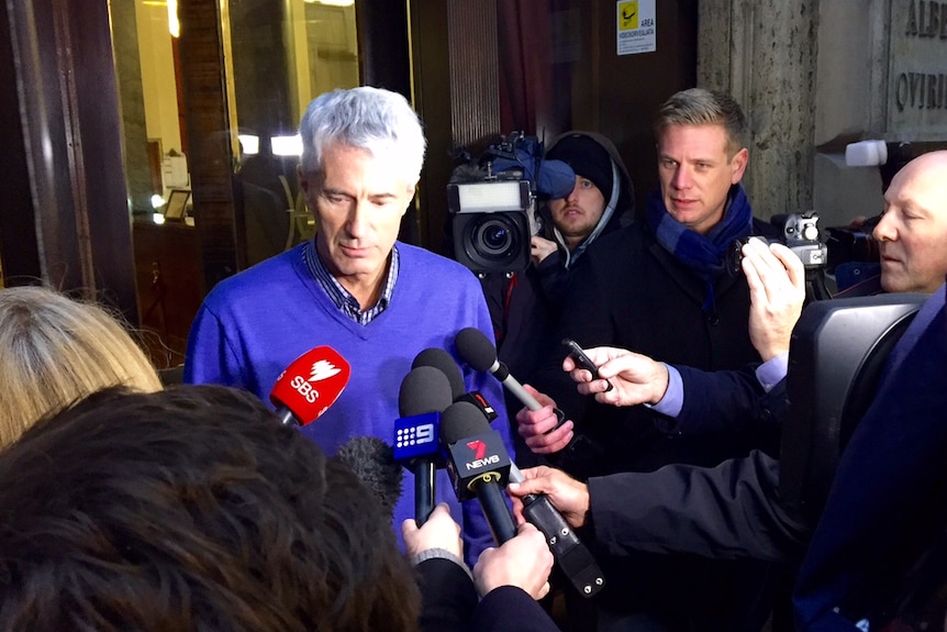 Anthony Foster speaks to several reporters on a street in Rome at night.