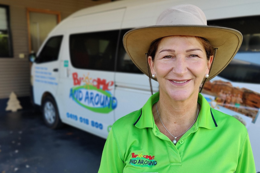 A woman wearing a bright green shirt and a wide-brim hat smiles for a photo standing in front of a minibus.