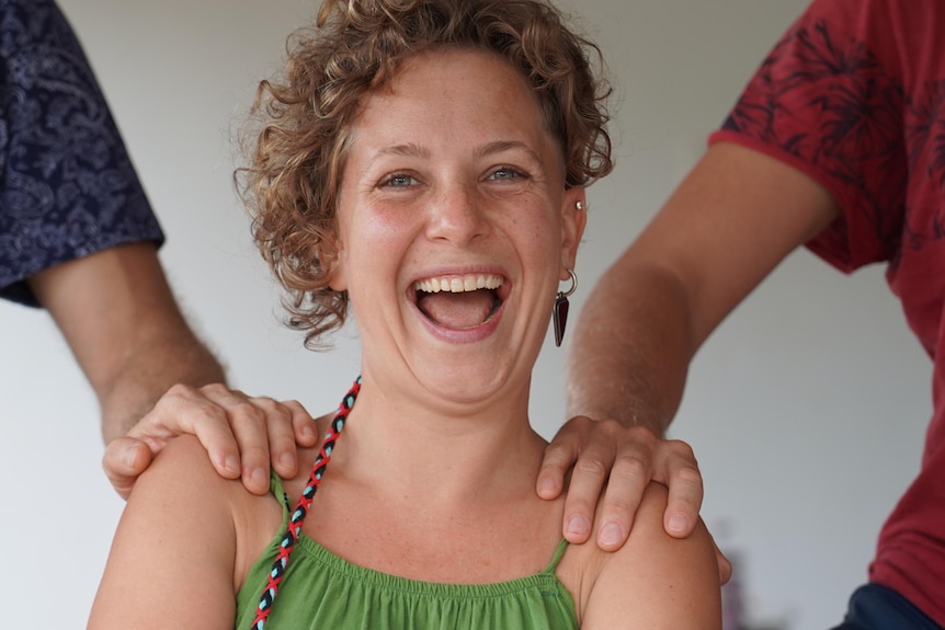 A young woman with curly hair laughs as two men place their hands on her shoulders