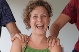 A young woman with curly hair laughs as two men place their hands on her shoulders