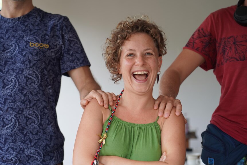 A curly-haired young woman laughs as two men put their hands on her shoulders