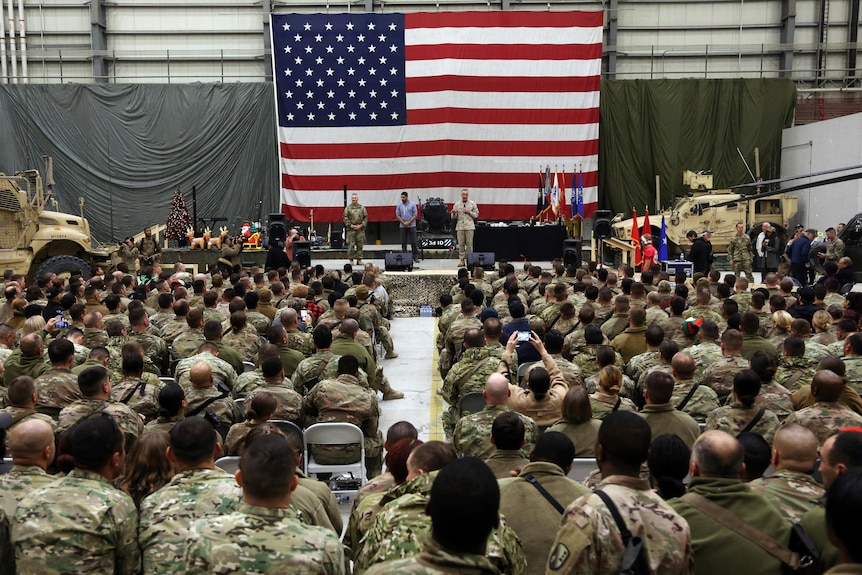 A group of US troops sit in a hanger watching a general give an address with a large American flag hanging behind him.
