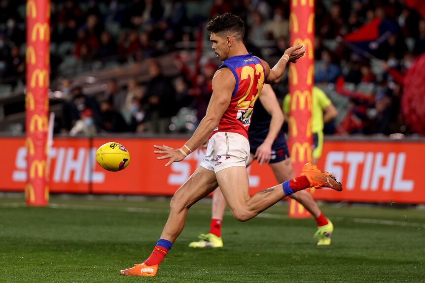 An Indigenous AFL footballer looks down as he swings his leg toward the ball while facing away from the goalposts during a game.