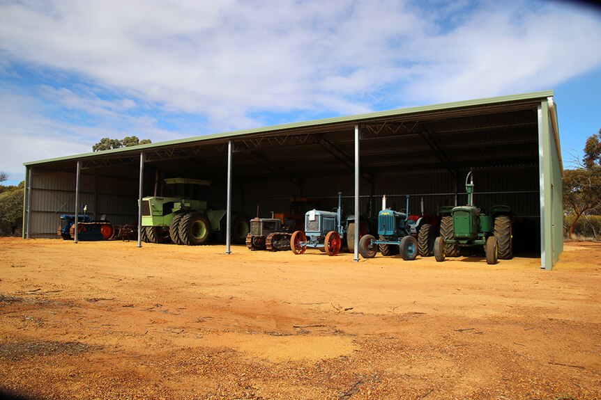 A line of colourful tractors sits in an open shed on the red dirt.
