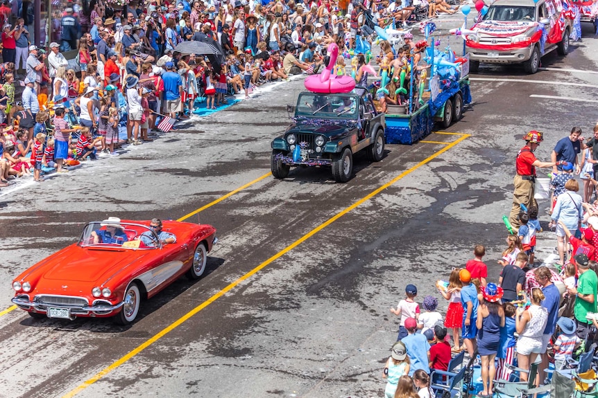 Onlookers watch a parade of colourful care decked out in American flag paraphernalia and people drive down a street