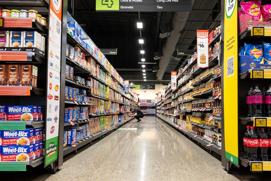 Looking down a near empty aisle of a supermarket. In the distance a staff member squats looking at a shelf.