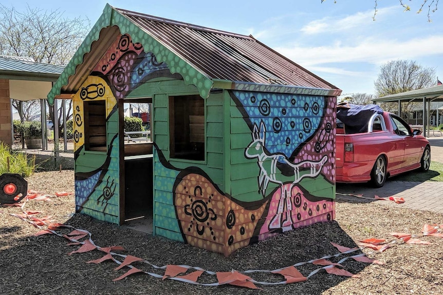 A small cubby house sitting on bark at a school has a kangaroo painted on it.