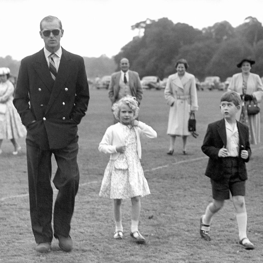 A black and white photo of Prince Philip wearing sunglasses and walking on grass next to his young daughter and son.