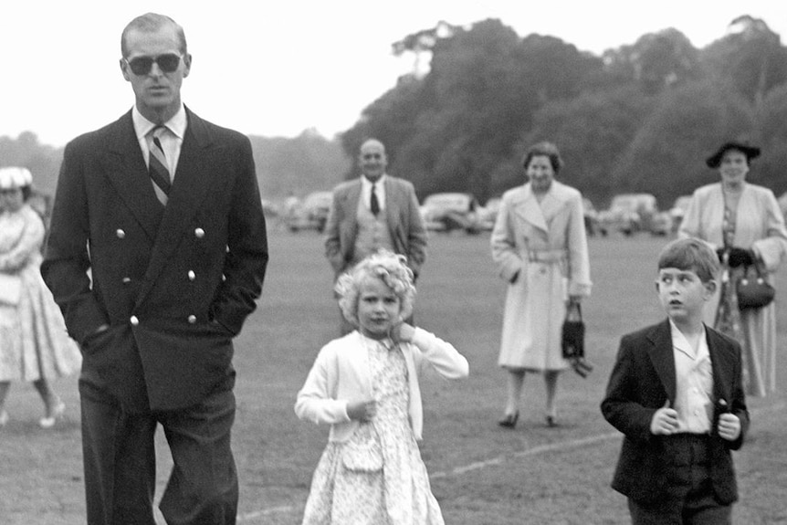 A black and white photo of Prince Philip wearing sunglasses and walking on grass next to his young daughter and son.